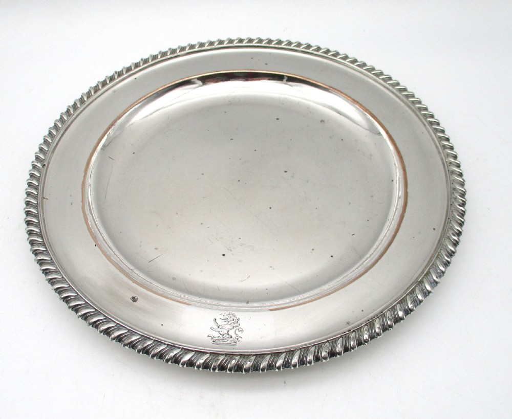 A circular Georgian Old Sheffield silver-plated dinner plate featuring a gadrooned rope-twist rim and an engraved armorial crest on the edge.