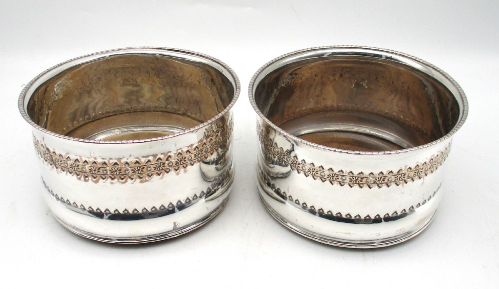 A pair of deep, circular silver-plated wine coasters featuring pierced floral decorative bands, beaded rims, and inset wooden bases.