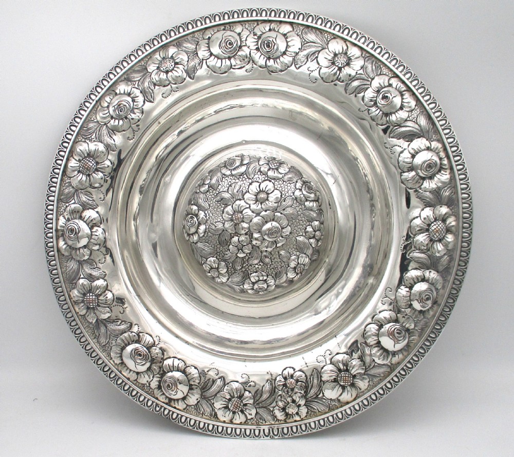 A circular silver-plated antique charger featuring an embossed floral rim and a raised central dome adorned with floral relief patterns.