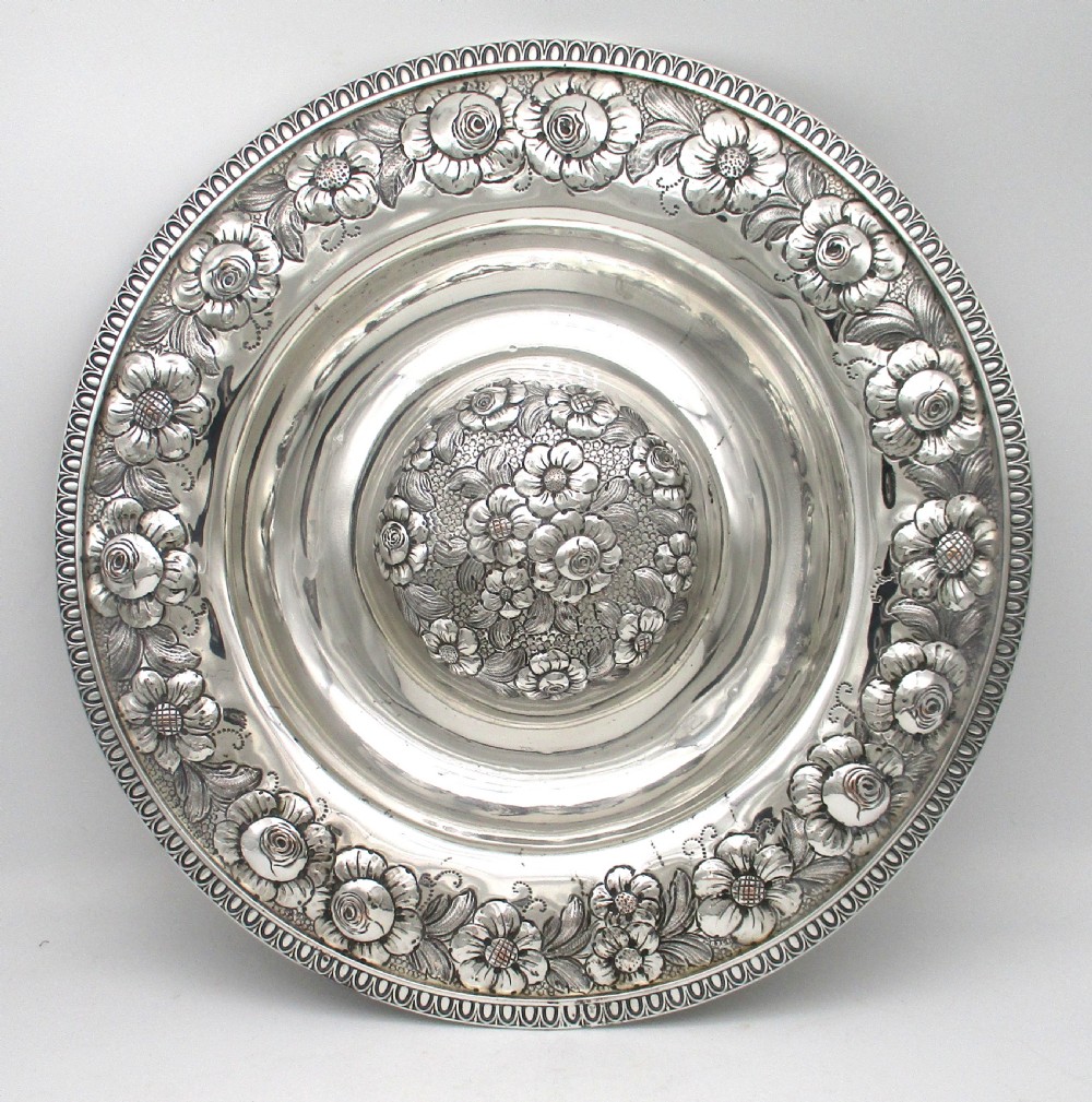 A circular silver-plated antique charger featuring an embossed floral rim and a raised central dome adorned with floral relief patterns. View II