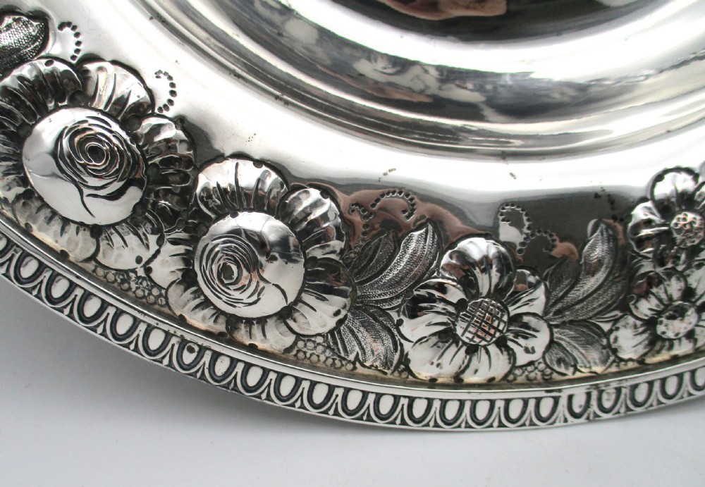 A circular silver-plated antique charger featuring an embossed floral rim and a raised central dome adorned with floral relief patterns. View IV