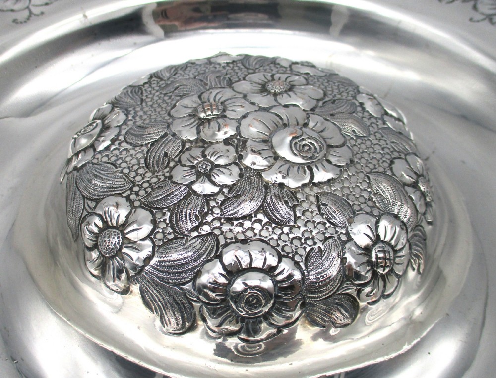 A circular silver-plated antique charger featuring an embossed floral rim and a raised central dome adorned with floral relief patterns. View V