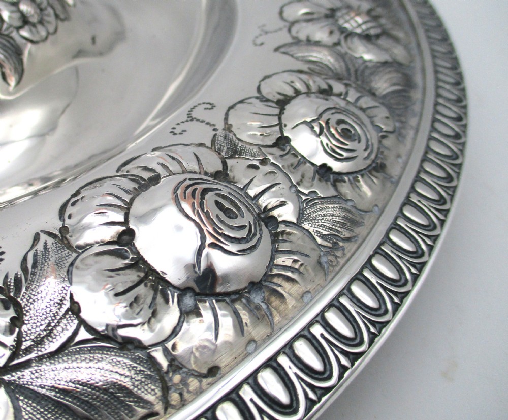 A circular silver-plated antique charger featuring an embossed floral rim and a raised central dome adorned with floral relief patterns. View VI