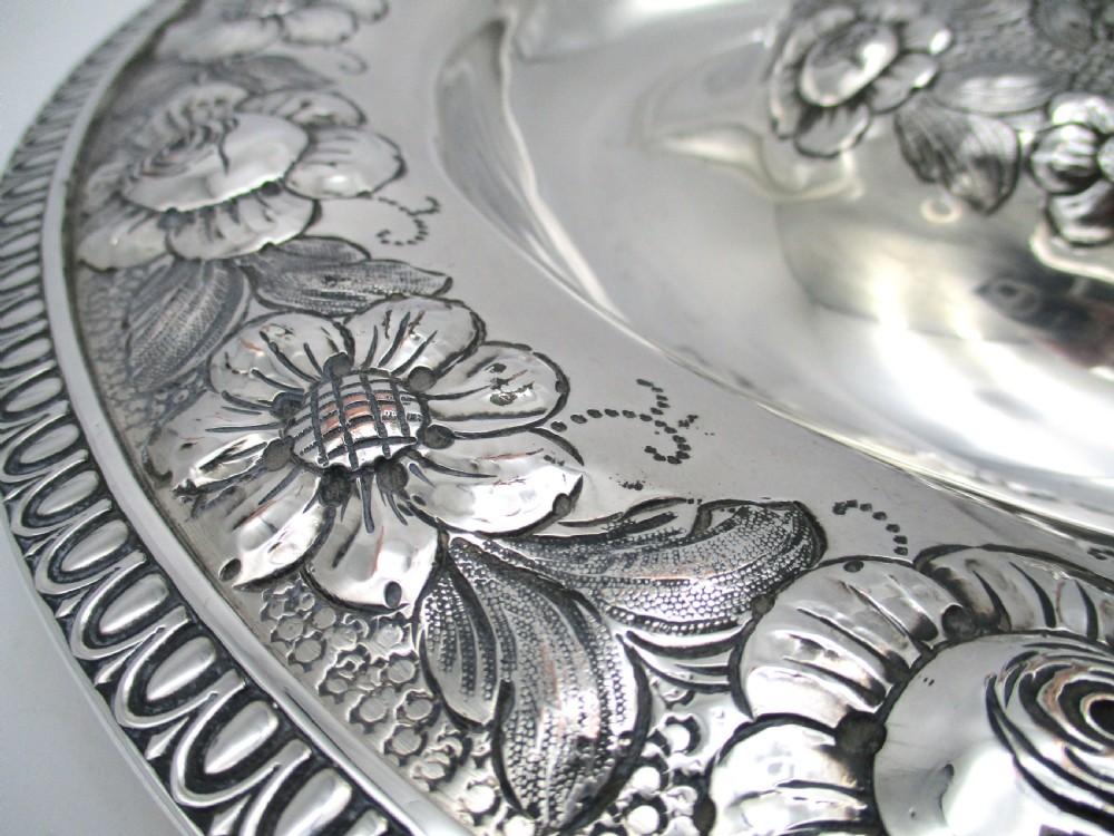A circular silver-plated antique charger featuring an embossed floral rim and a raised central dome adorned with floral relief patterns. View VII