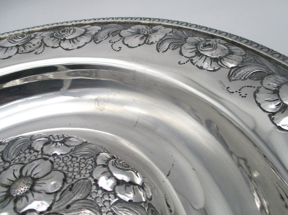 A circular silver-plated antique charger featuring an embossed floral rim and a raised central dome adorned with floral relief patterns. View VIII