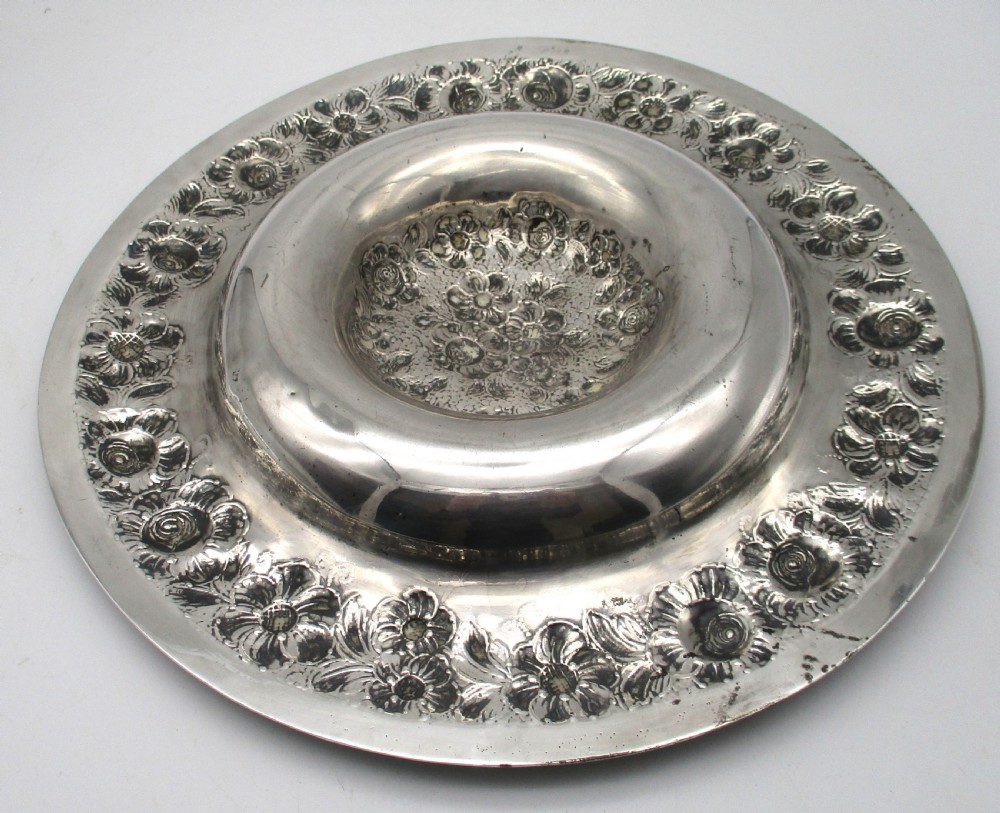 A circular silver-plated antique charger featuring an embossed floral rim and a raised central dome adorned with floral relief patterns. View IX