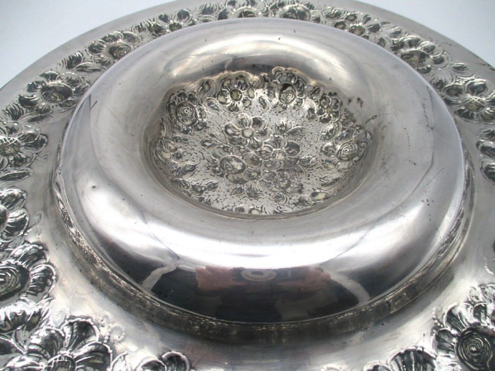A circular silver-plated antique charger featuring an embossed floral rim and a raised central dome adorned with floral relief patterns. View X