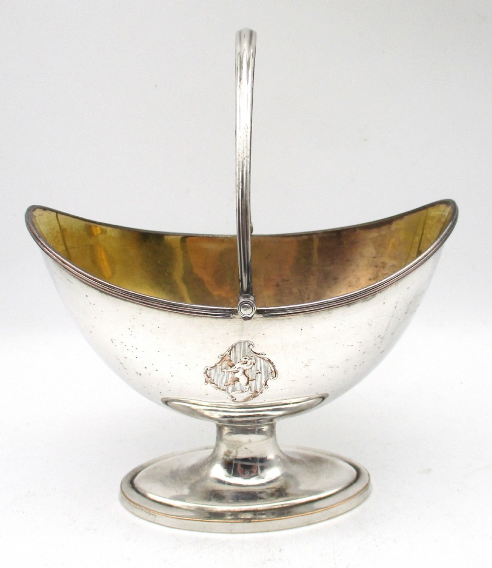 An eighteenth-century boat-shaped silver plated sweetmeat basket featuring a pedestal base, swing handle, heraldic crest engraving, and a gilded interior.