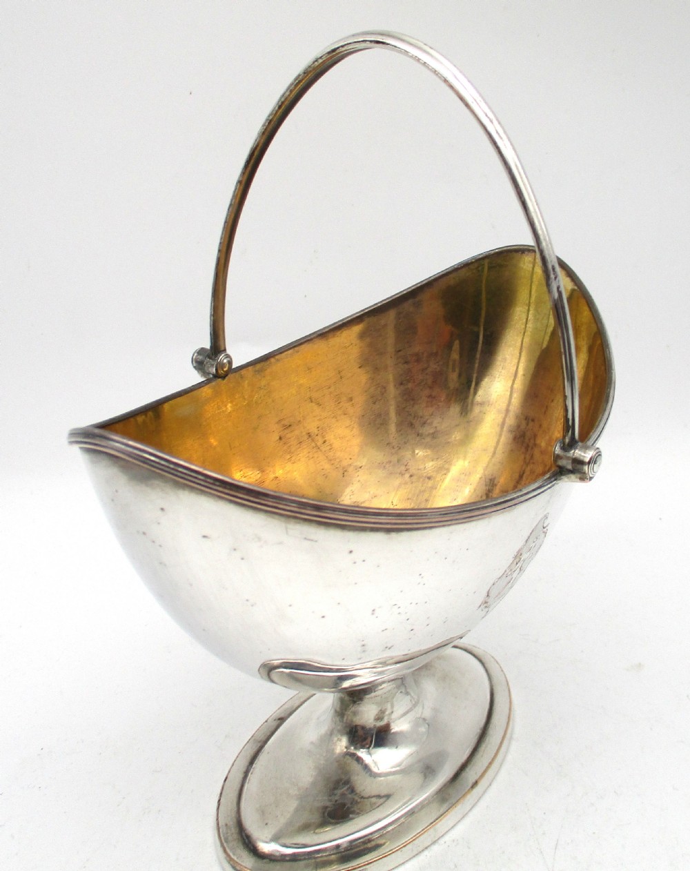 An eighteenth-century boat-shaped silver plated sweetmeat basket featuring a pedestal base, swing handle, heraldic crest engraving, and a gilded interior. View V