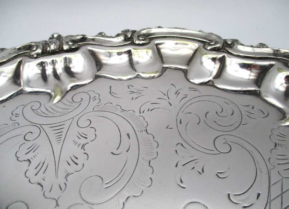 A circular Victorian silver-plated salver featuring an engraved floral scroll centre, shell-and-scroll moulded border, and short cast feet. View VI