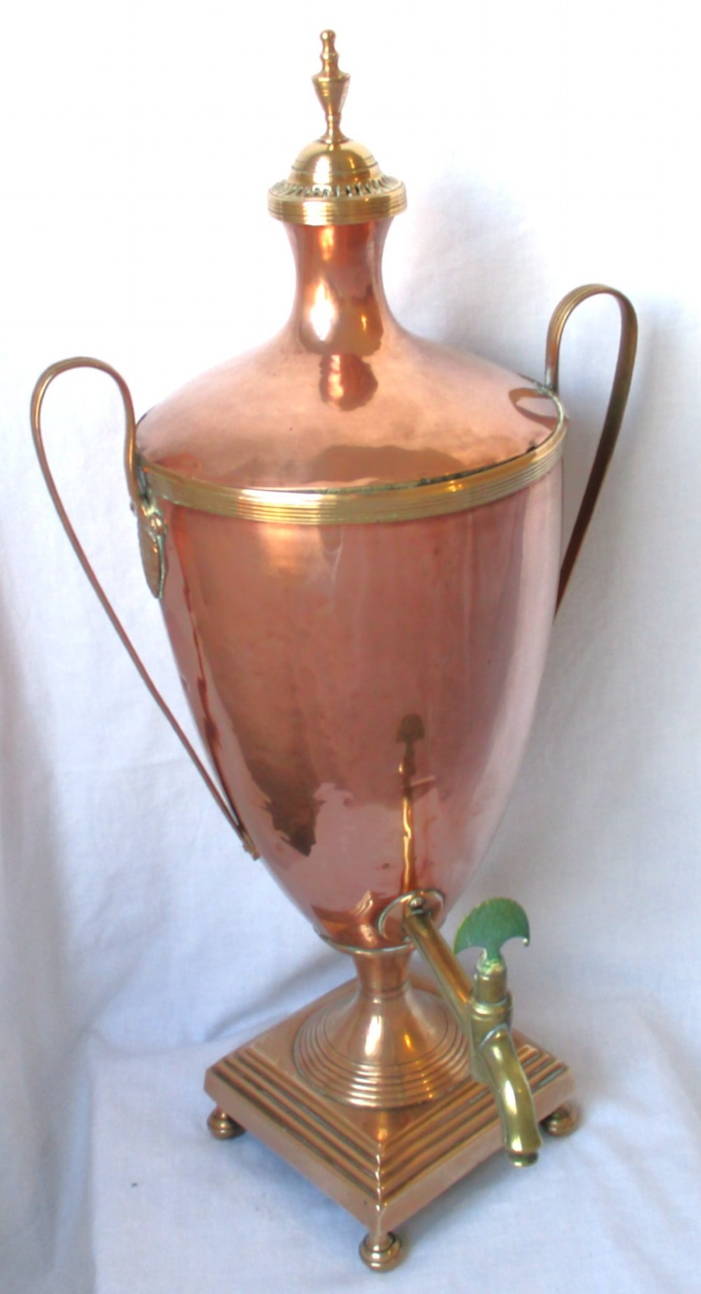 An early nineteenth-century copper tea urn featuring loop handles, a brass spigot, a decorative finial, and a square stepped pedestal base.