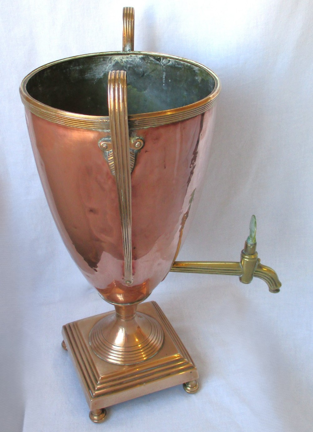 An early nineteenth-century copper tea urn featuring loop handles, a brass spigot, a decorative finial, and a square stepped pedestal base. View VIII