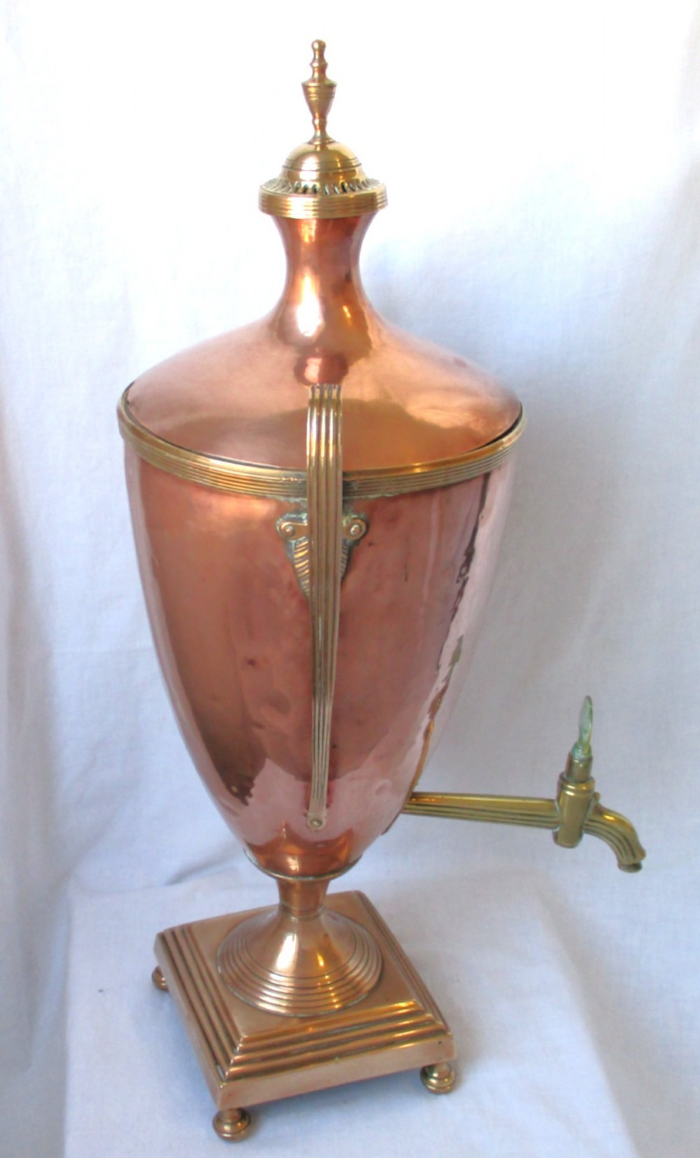 An early nineteenth-century copper tea urn featuring loop handles, a brass spigot, a decorative finial, and a square stepped pedestal base. View IX