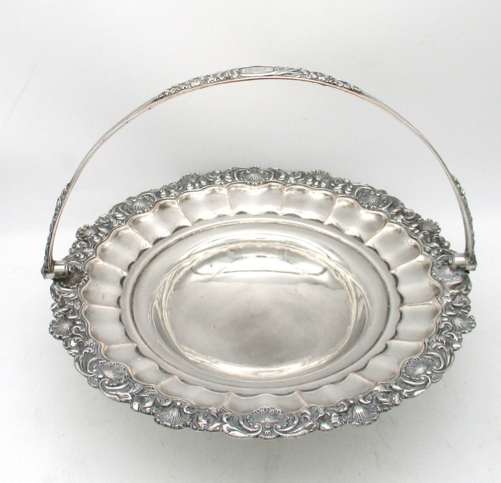 A round Old Sheffield Plate silver basket with a fluted rim, shell border motifs, an engraved crest, and a overhead swing handle.