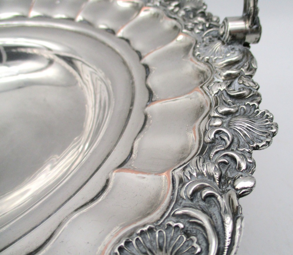 A round Old Sheffield Plate silver basket with a fluted rim, shell border motifs, an engraved crest, and a overhead swing handle. View IV