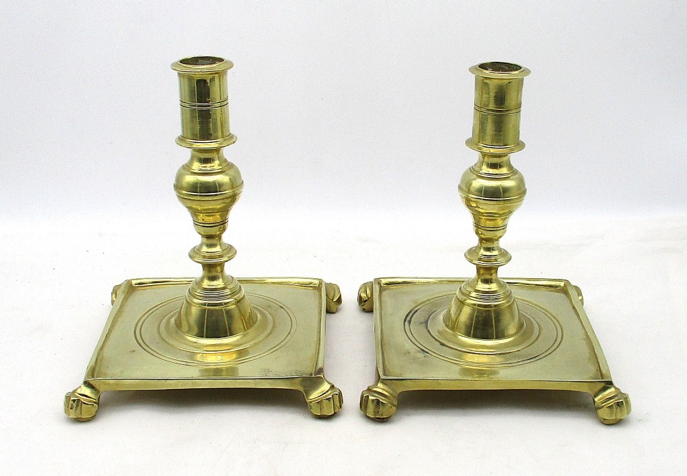 A pair of polished brass candlesticks with turned baluster stems and square bases featuring corner feet.