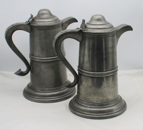 A pair of matching 19th-century pewter communion flagons featuring domed hinged lids, thumbpiece levers, scroll handles, and engraved inscriptions on the cylindrical bodies.