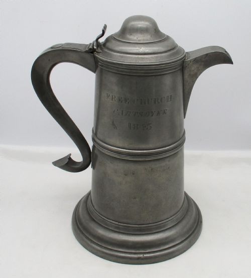 A pair of matching 19th-century pewter communion flagons featuring domed hinged lids, thumbpiece levers, scroll handles, and engraved inscriptions on the cylindrical bodies. View II
