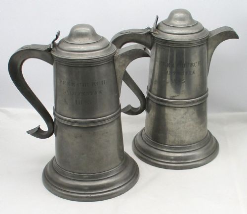 A pair of matching 19th-century pewter communion flagons featuring domed hinged lids, thumbpiece levers, scroll handles, and engraved inscriptions on the cylindrical bodies. View XII