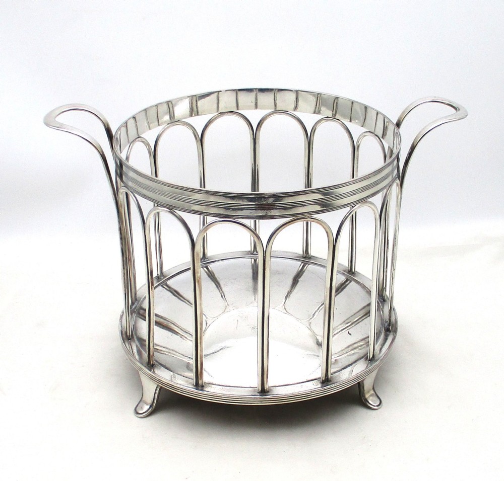 A silver-plated cylindrical bottle coaster featuring arched wirework sides, twin looped handles, a reeded rim, and small curved feet.