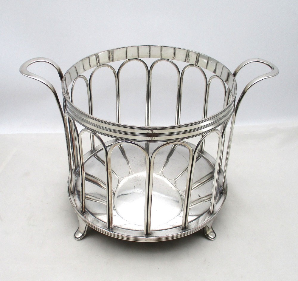 A silver-plated cylindrical bottle coaster featuring arched wirework sides, twin looped handles, a reeded rim, and small curved feet. View II