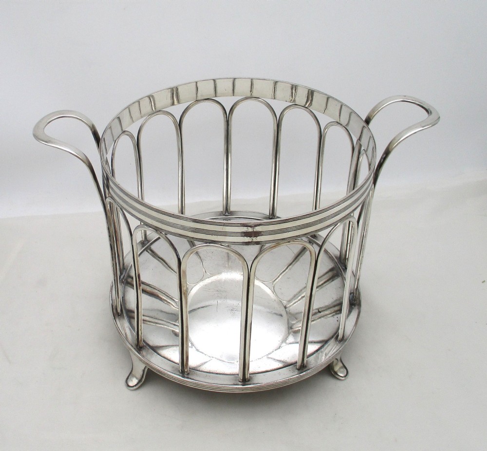 A silver-plated cylindrical bottle coaster featuring arched wirework sides, twin looped handles, a reeded rim, and small curved feet. View IV