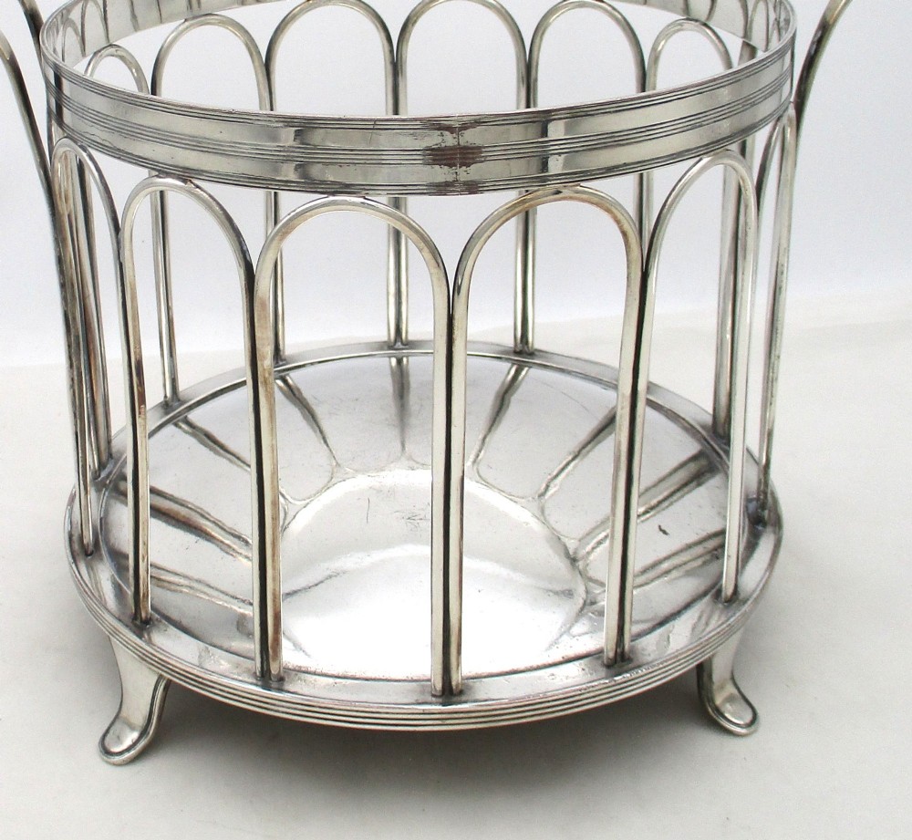 A silver-plated cylindrical bottle coaster featuring arched wirework sides, twin looped handles, a reeded rim, and small curved feet. View V