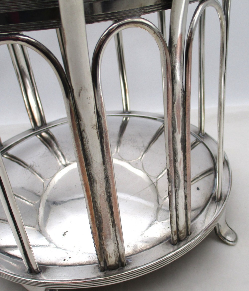 A silver-plated cylindrical bottle coaster featuring arched wirework sides, twin looped handles, a reeded rim, and small curved feet. View VI