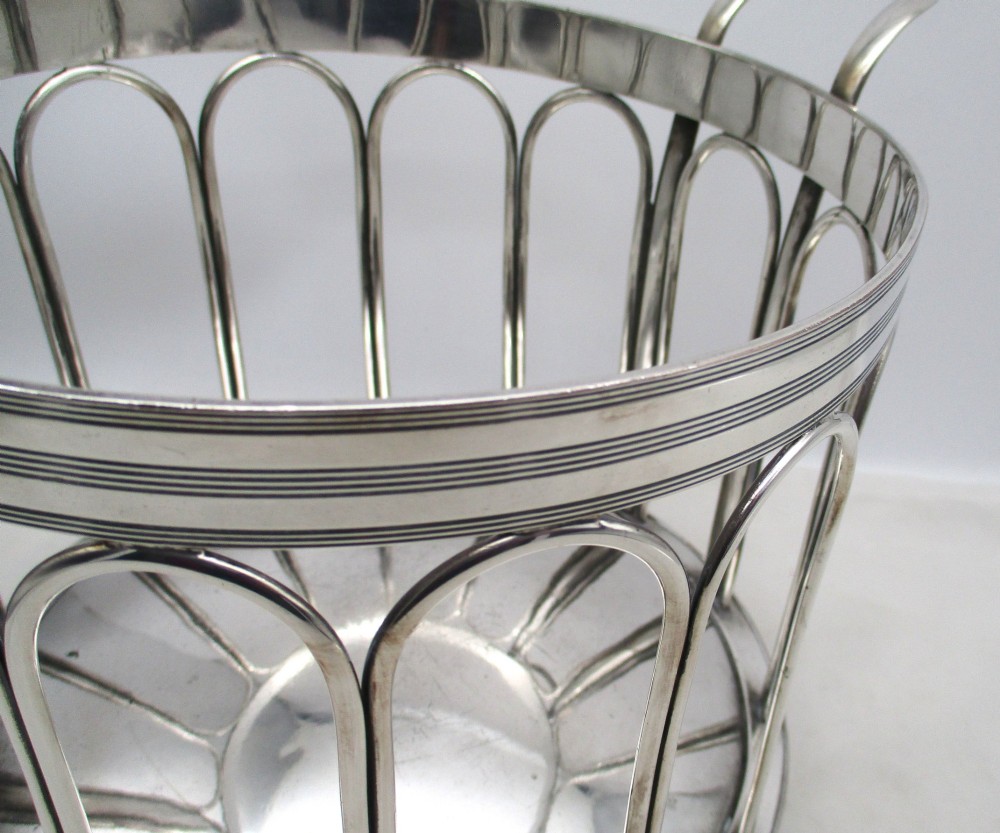 A silver-plated cylindrical bottle coaster featuring arched wirework sides, twin looped handles, a reeded rim, and small curved feet. View VII