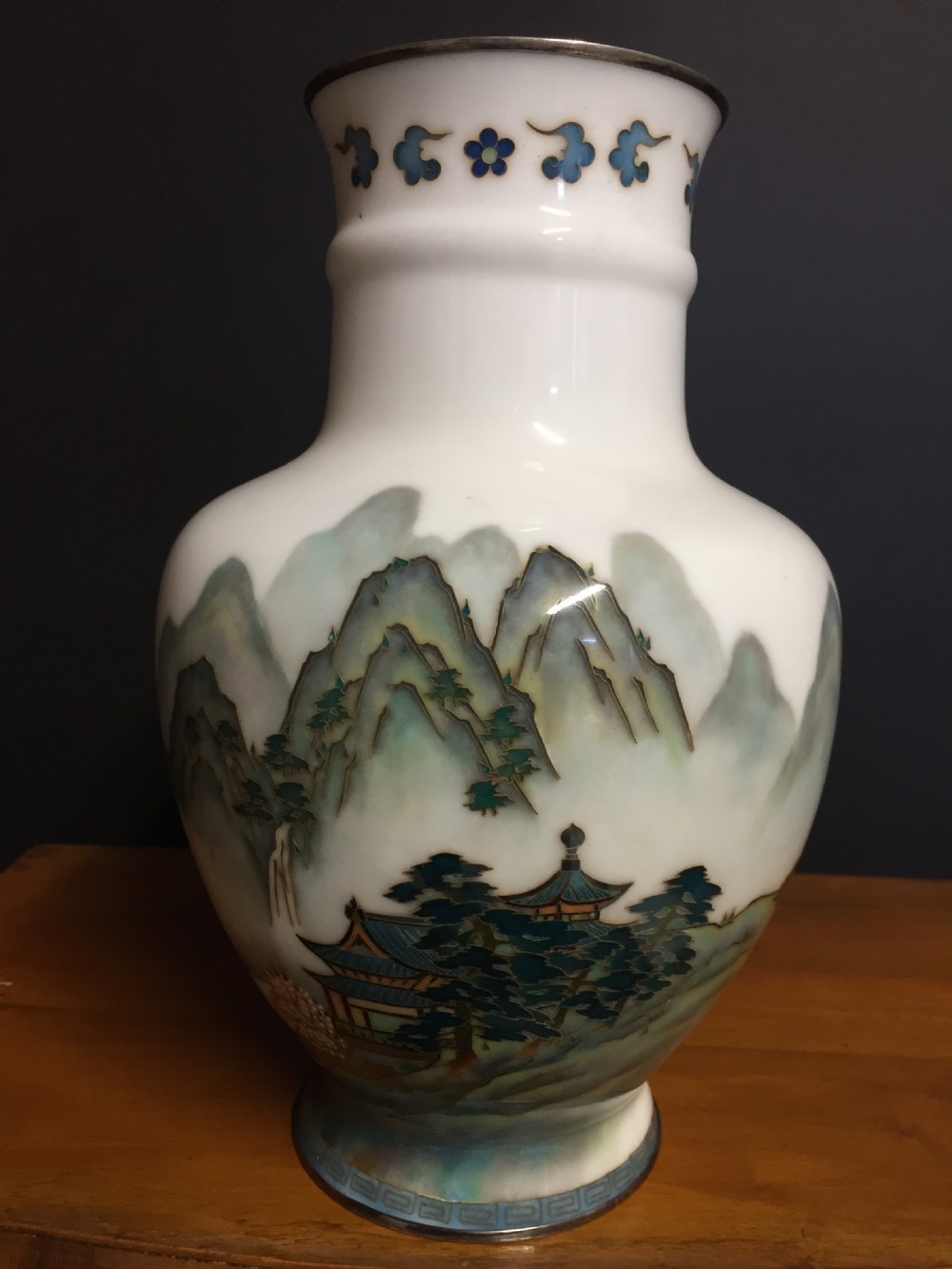 A white Japanese cloisonne enamel vase depicting a green and blue landscape with mountains, pagodas, and trees, finished with a silver rim.