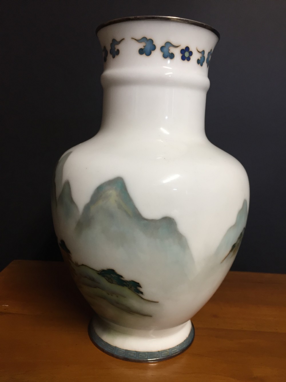 A white Japanese cloisonne enamel vase depicting a green and blue landscape with mountains, pagodas, and trees, finished with a silver rim. View III