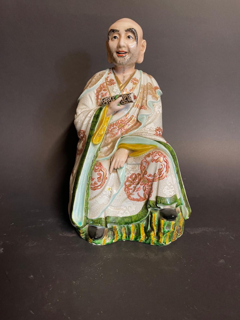 Japanese porcelain figure depicting a seated monk holding a scroll, dressed in white, green, and iron-red patterned robes.