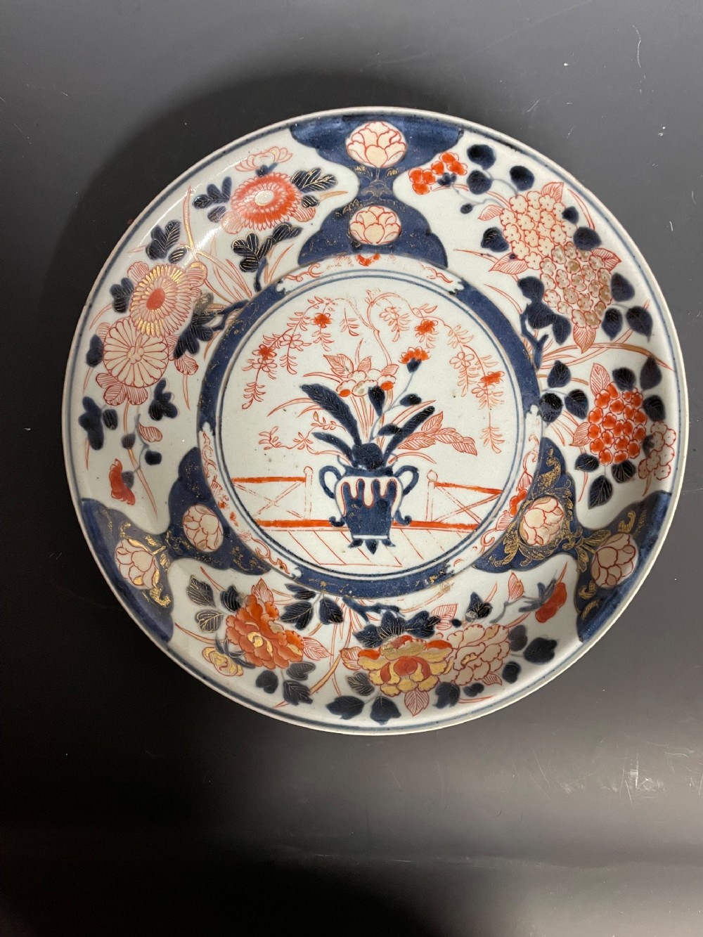 A circular Japanese Arita porcelain dish decorated with cobalt blue underglaze, iron red enamels, and gold leaf floral patterns surrounding a central urn motif.