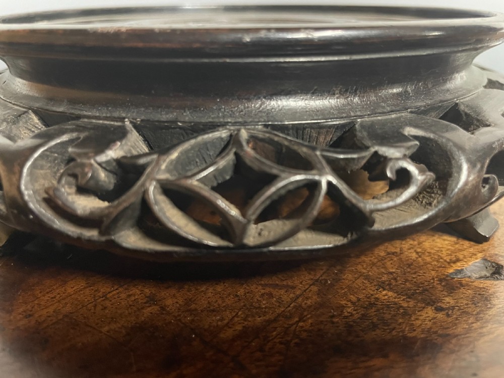 A circular dark hardwood vase stand featuring hand-carved openwork details around the apron and a recessed top surface. View VI
