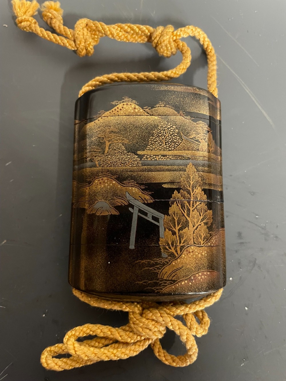 A small Japanese multi-tiered black lacquer inro box decorated with gold maki-e landscape imagery and suspended by a woven yellow cord.