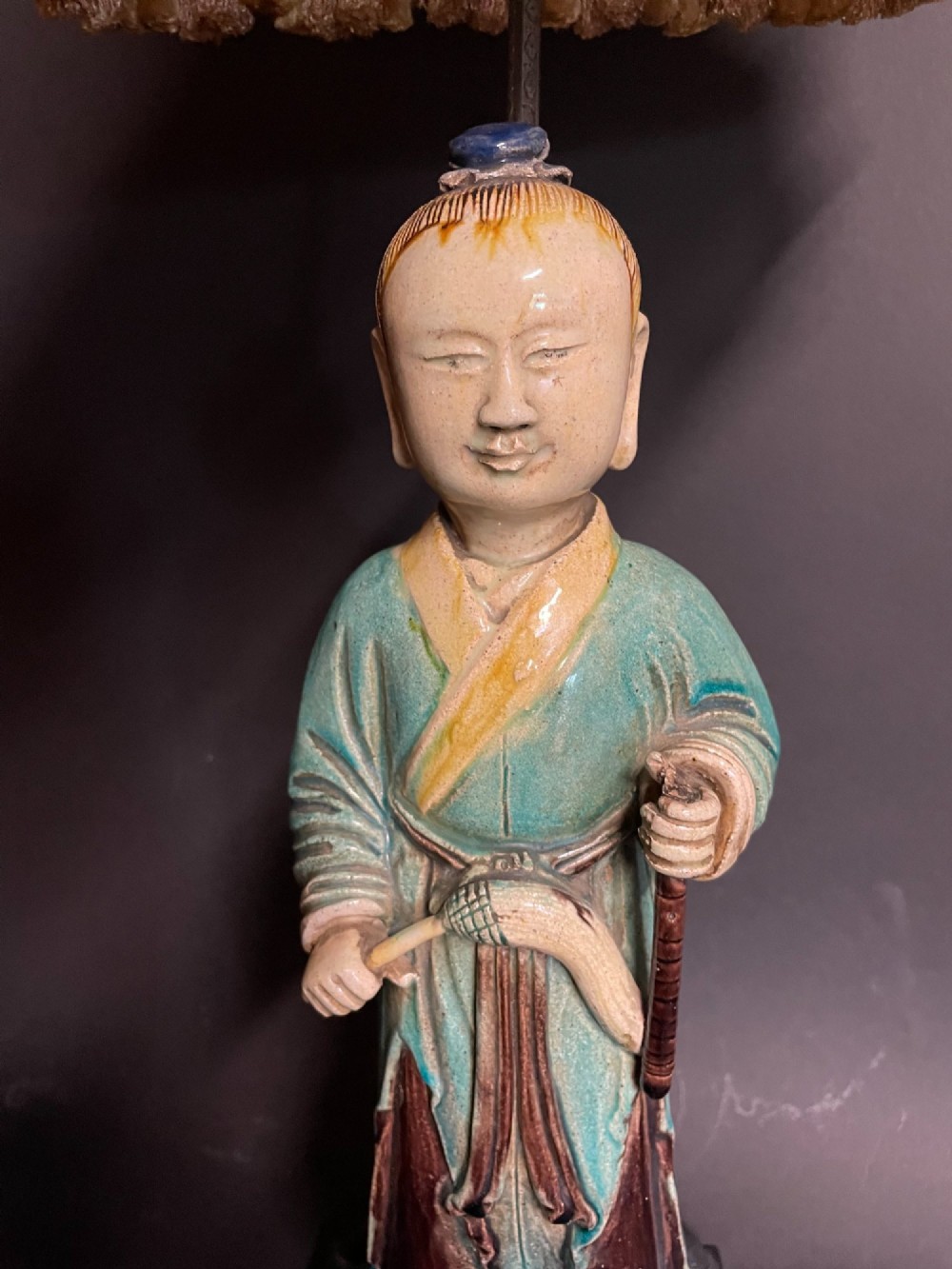 A Chinese porcelain figure table lamp featuring a green and brown glazed figurine on a black base topped with a tasseled fabric shade showing a painted landscape. View IV