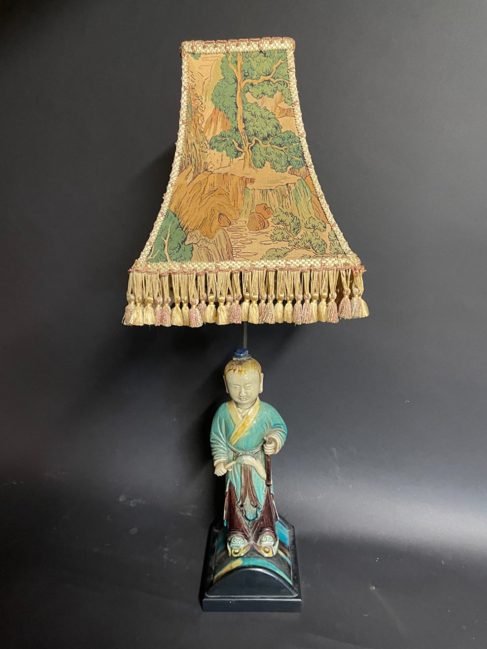 A Chinese porcelain figure table lamp featuring a green and brown glazed figurine on a black base topped with a tasseled fabric shade showing a painted landscape.
