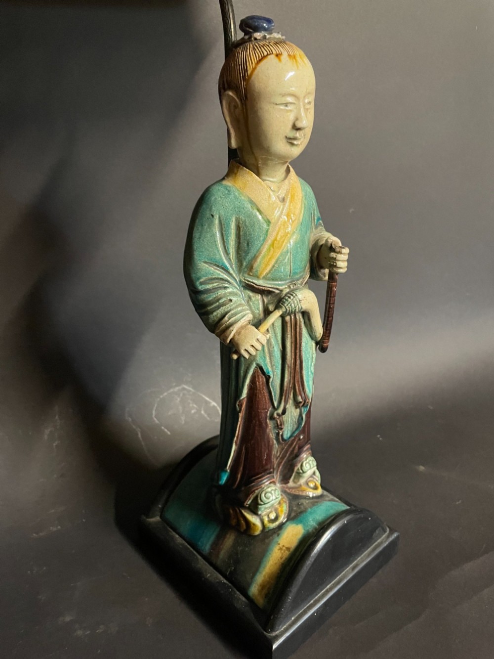 A Chinese porcelain figure table lamp featuring a green and brown glazed figurine on a black base topped with a tasseled fabric shade showing a painted landscape. View V