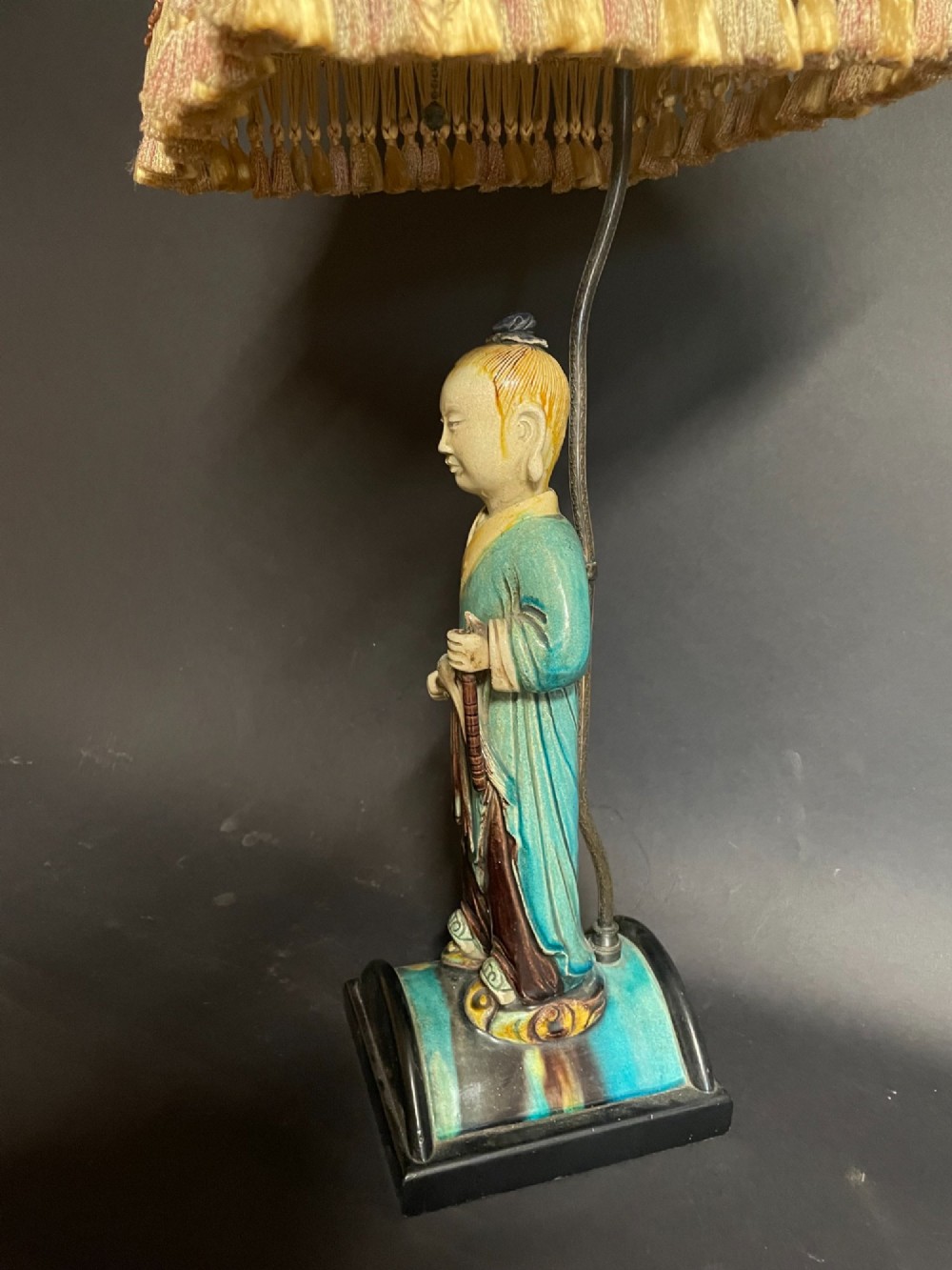 A Chinese porcelain figure table lamp featuring a green and brown glazed figurine on a black base topped with a tasseled fabric shade showing a painted landscape. View VII