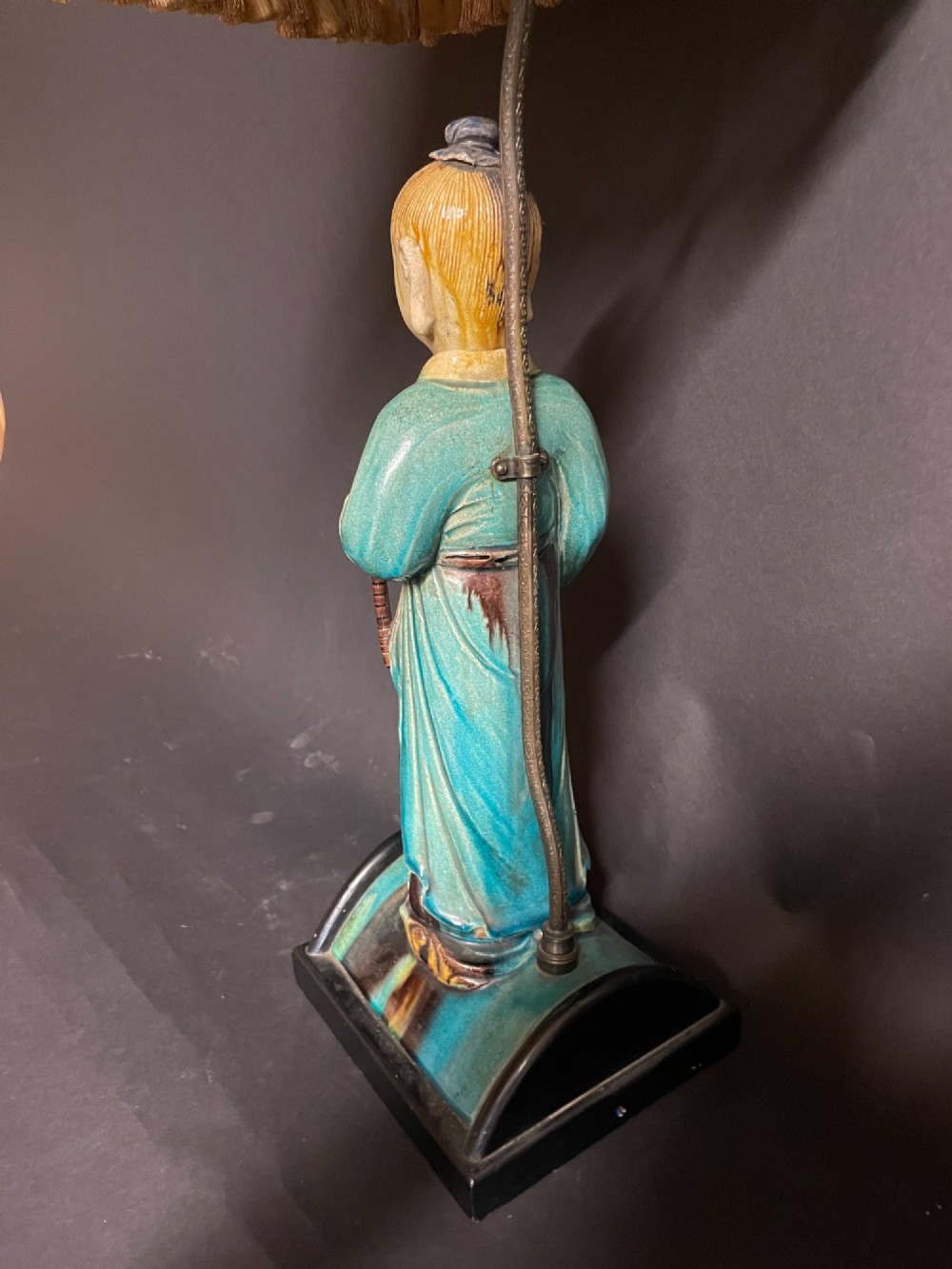 A Chinese porcelain figure table lamp featuring a green and brown glazed figurine on a black base topped with a tasseled fabric shade showing a painted landscape. View IX