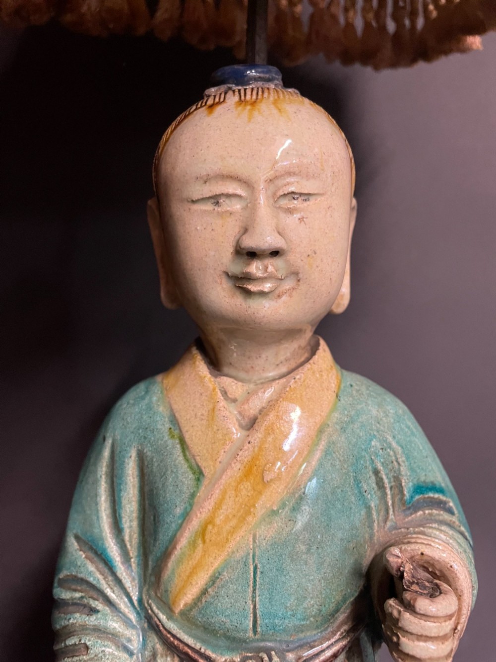 A Chinese porcelain figure table lamp featuring a green and brown glazed figurine on a black base topped with a tasseled fabric shade showing a painted landscape. View XI
