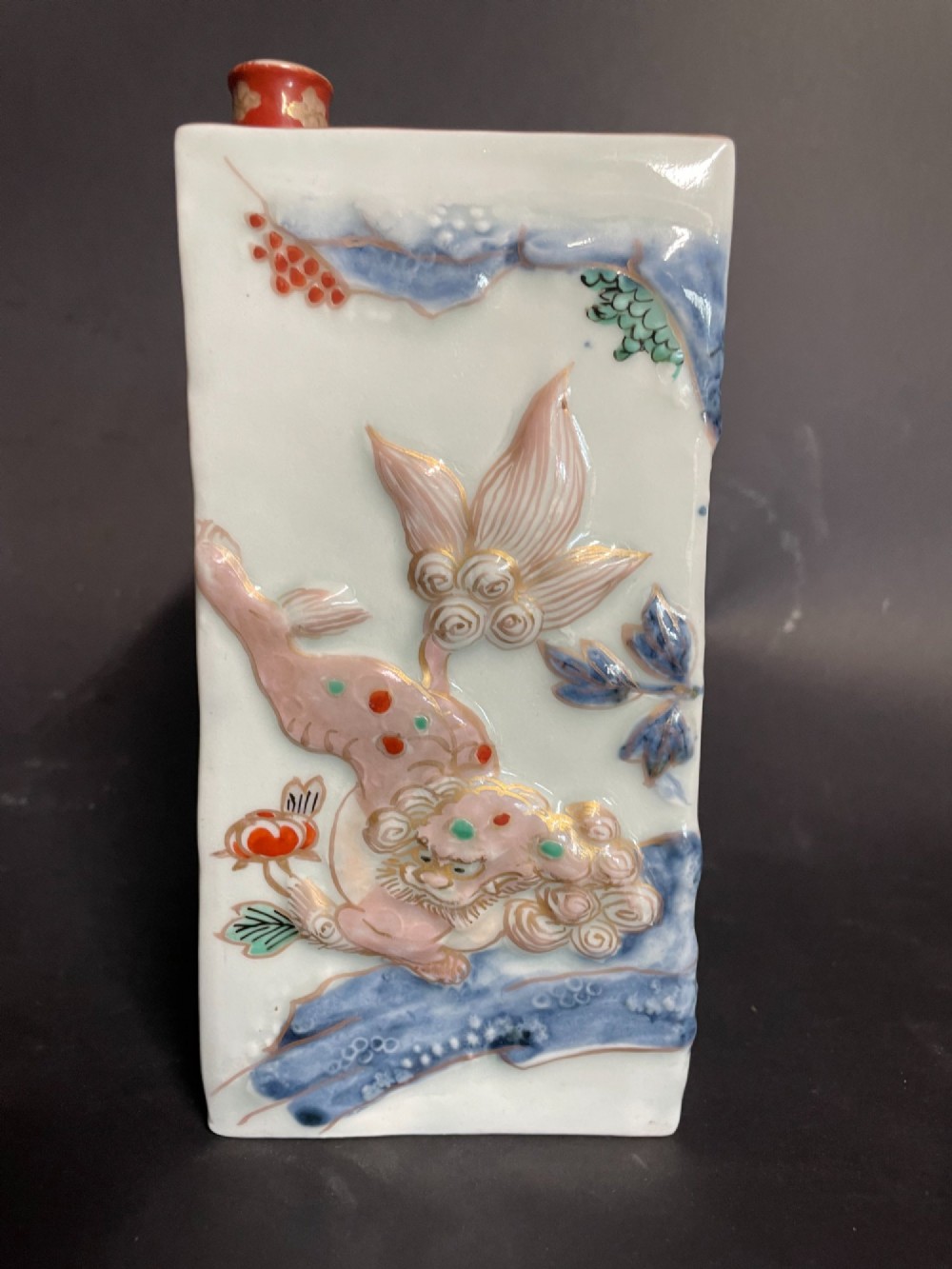 A rectangular off-white Japanese porcelain tokuri flask featuring relief carvings and hand-painted polychrome enamel depictions of a Shishi lion dog and floral foliage. View V