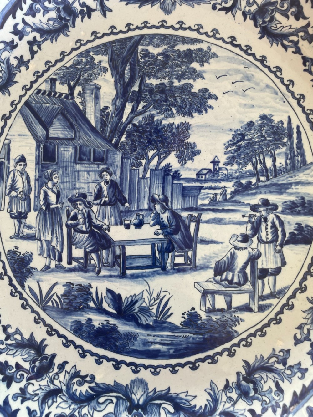 A circular Dutch Delft tin-glazed earthenware charger decorated in cobalt blue and white with a central pictorial tavern scene surrounded by ornate floral and cartouche borders. View II