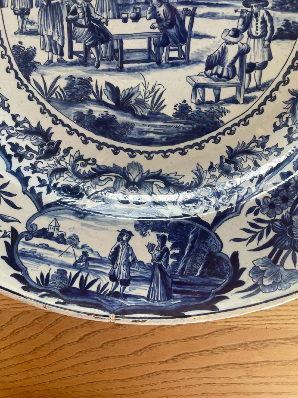 A circular Dutch Delft tin-glazed earthenware charger decorated in cobalt blue and white with a central pictorial tavern scene surrounded by ornate floral and cartouche borders. View V