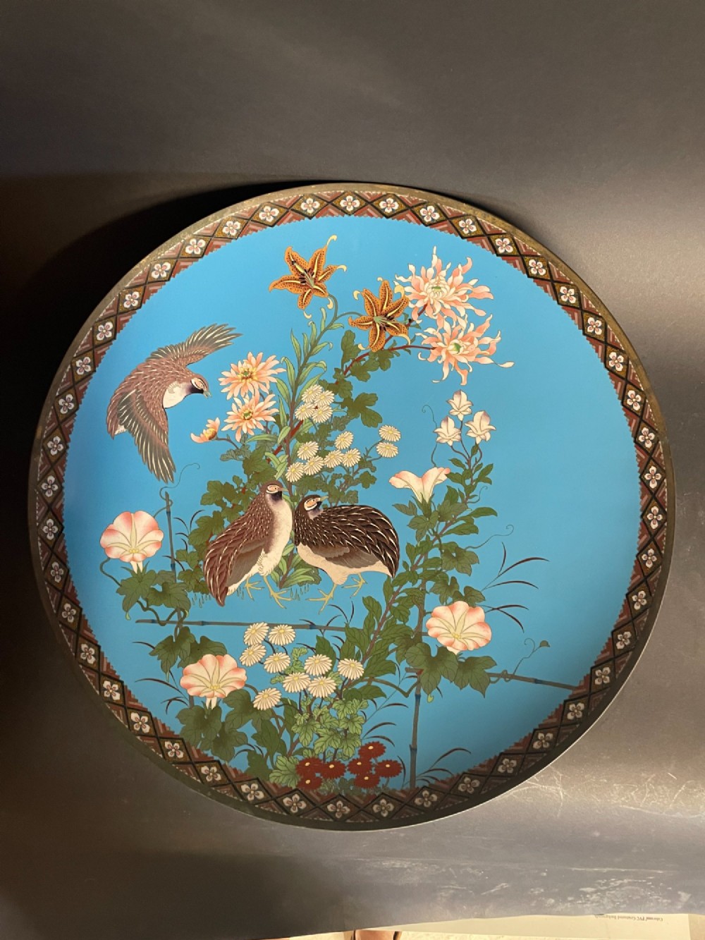 A circular Japanese cloisonne enamel charger depicting quails among chrysanthemums and lilies against a sky blue background with a patterned geometric border.