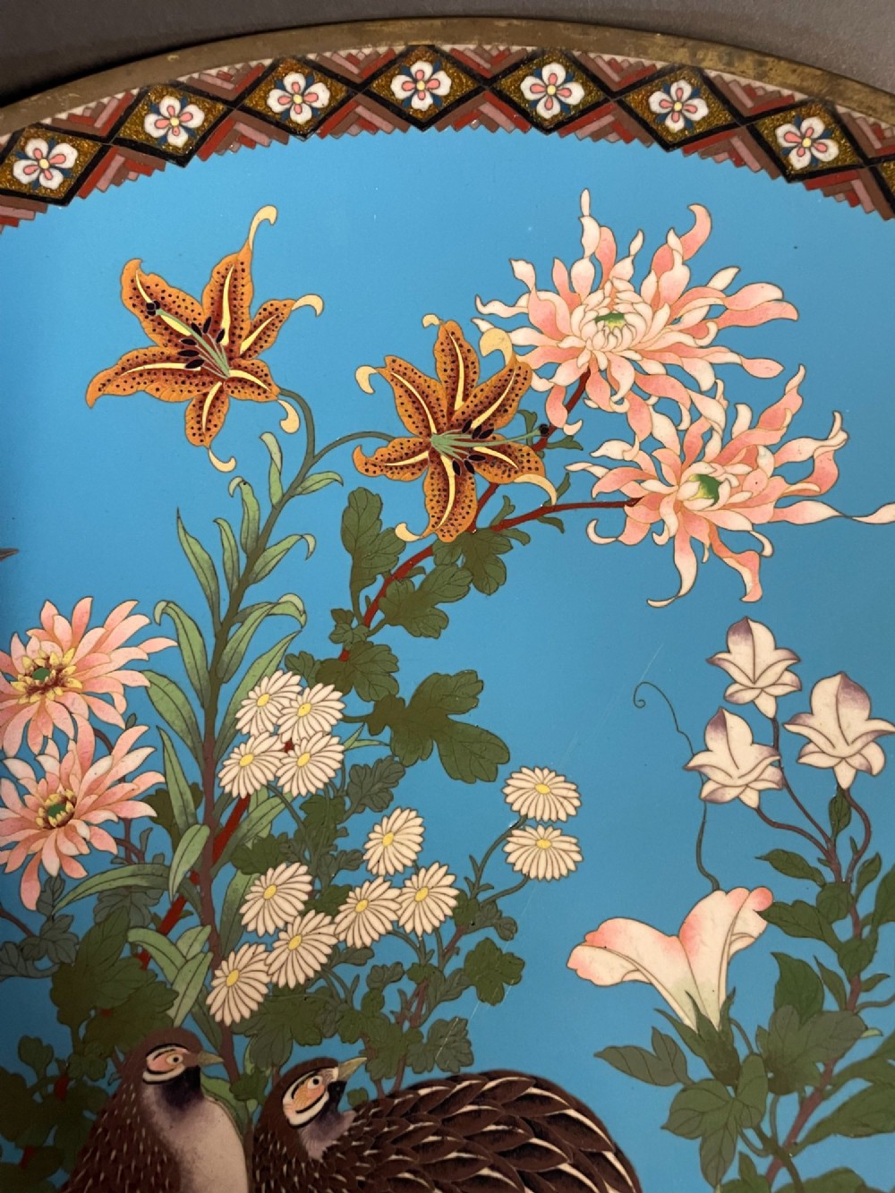 A circular Japanese cloisonne enamel charger depicting quails among chrysanthemums and lilies against a sky blue background with a patterned geometric border. View III