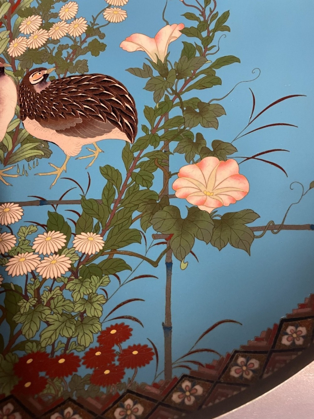 A circular Japanese cloisonne enamel charger depicting quails among chrysanthemums and lilies against a sky blue background with a patterned geometric border. View IV