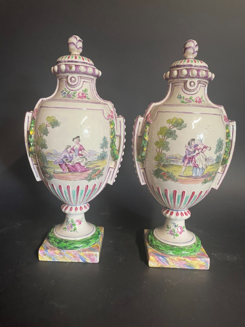 A pair of antique French Sceaux faience lidded urns decorated with hand-painted pastoral scenes, floral garlands, and moulding on square bases.