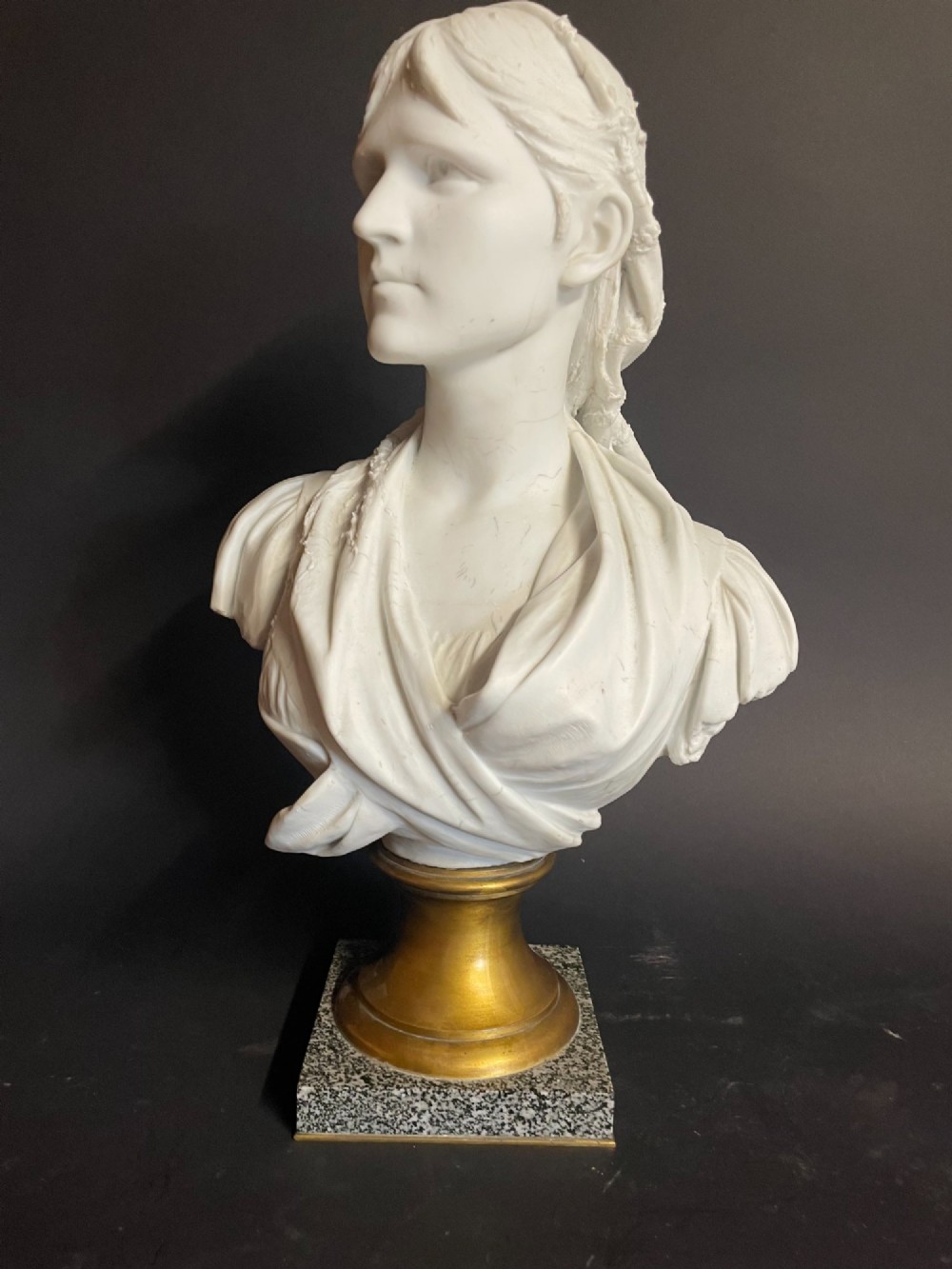 A white carved bust of a woman with draped classical clothing, mounted on a turned gold-tone socle and a square speckled granite base.