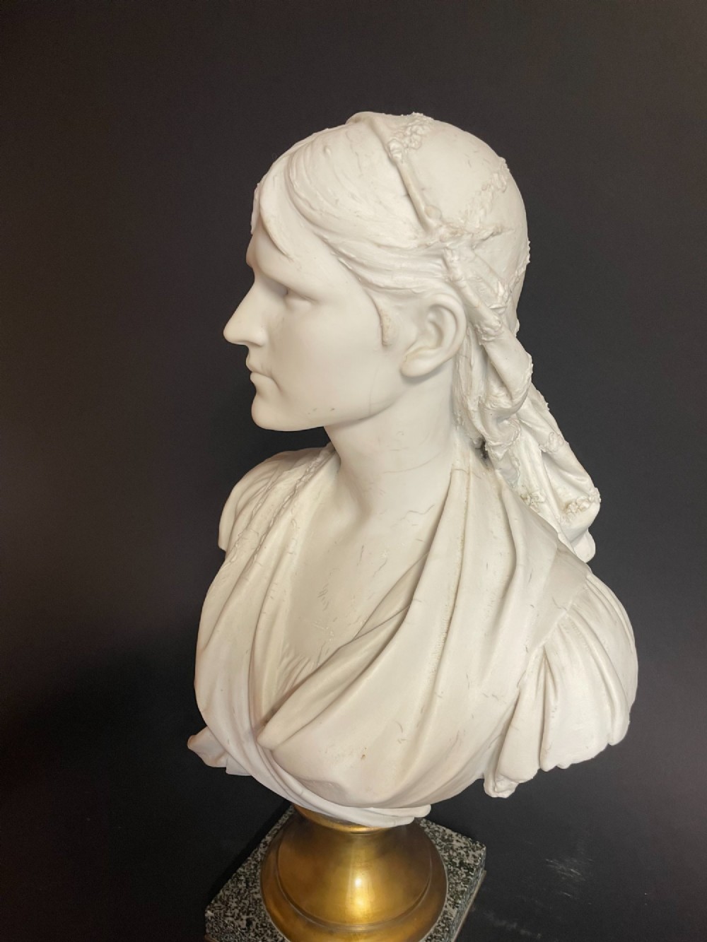 A white carved bust of a woman with draped classical clothing, mounted on a turned gold-tone socle and a square speckled granite base. View II